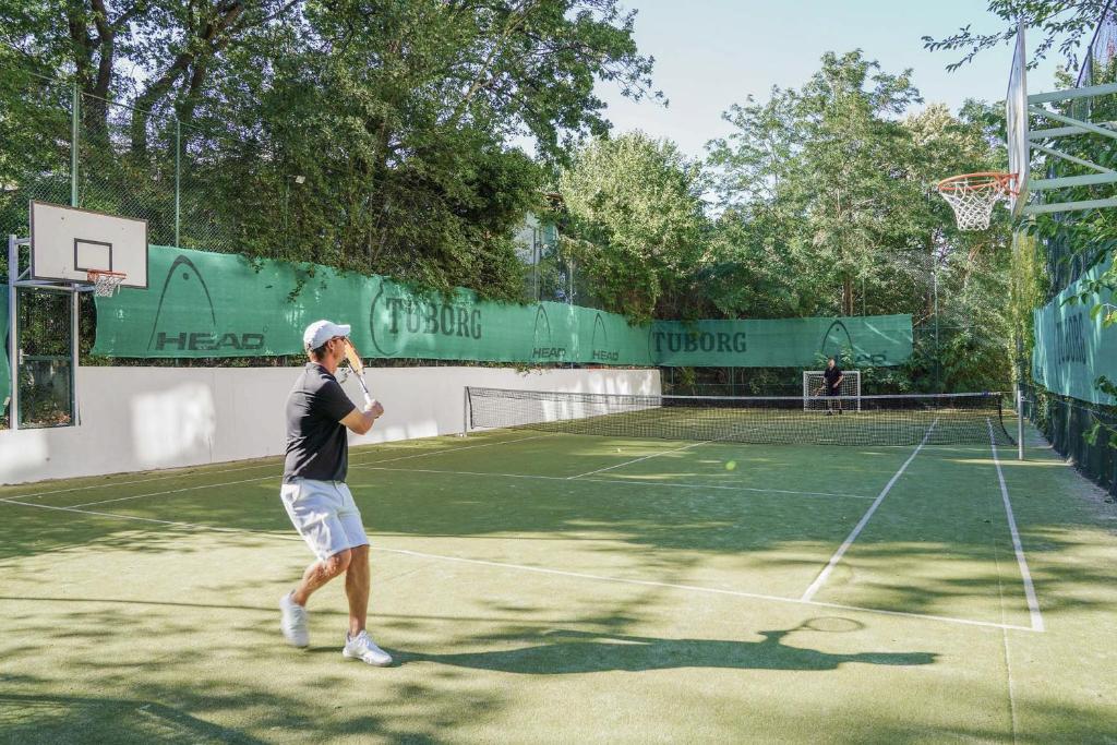 Tennis spēlētājs, kas sit bumbu uz zaļā laukuma, blakus basketbola grozs un tīkla laukums. Apkārt ir zaļi koki un reklāmas baneri.