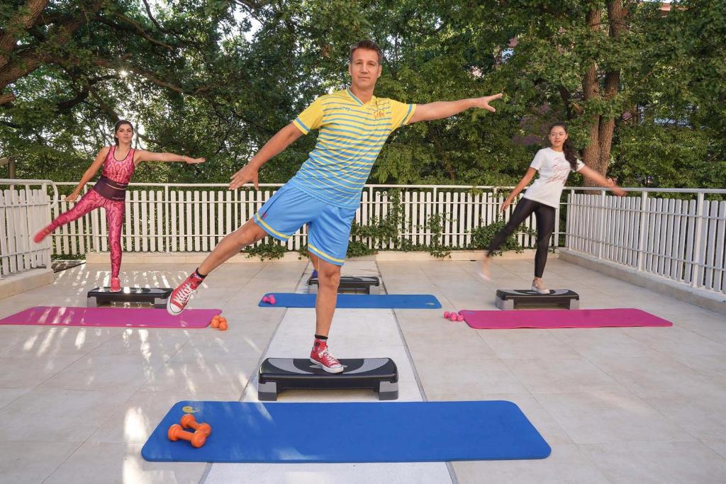 Grupas nodarbība uz terases, kur dalībnieki veic vingrojumus uz step platformām. Pievilcīga vide ar zaļu fonu un dažādu krāsu vingrošanas paklājiem.