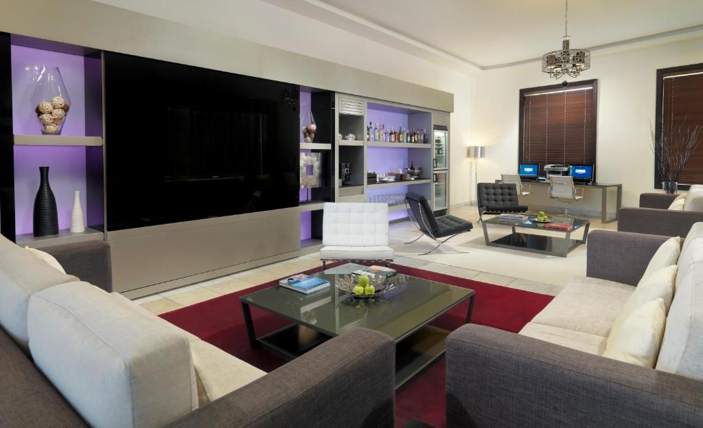 Mūsdienīgs viesistabas interjers ar elegantiem dīvāniem, stikla galdiem un modernu televizoru. Telpā ir arī darba zona un bāra plaukts, radot ērti pieejamu un stilīgu atmosfēru.