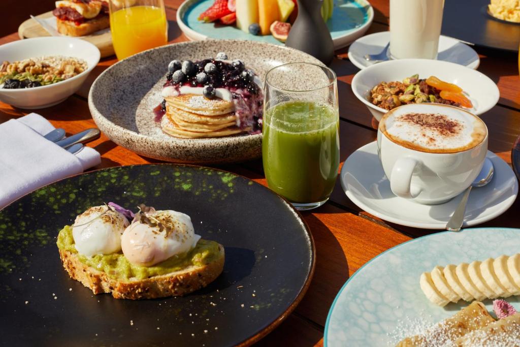 Pusdienu galda aina ar dažādiem ēdieniem, tostarp pankūkām ar ogām, avokado grauzdiņiem ar pošētiem olu dzeltenumiem un veselīgu dzērienu. Svaigi augļi un kafija papildina šo garšīgo brokastu piedāvājumu.