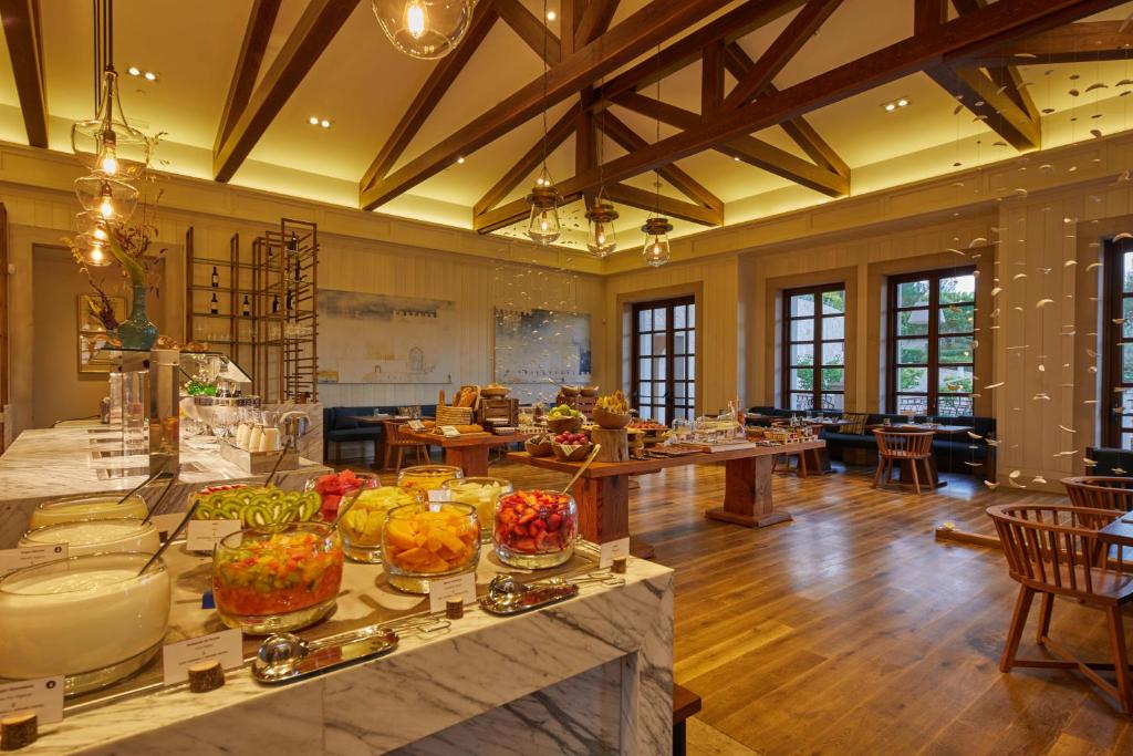 Labi aprīkots restorāns ar elegantu interjeru, kurā redzamas dažādas augļu un desertu galda izkārtojumi. Gaismas ķermeņi un koka konstrukcijas piešķir siltu atmosfēru.