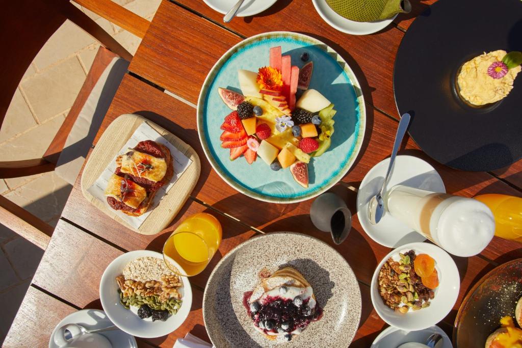 Krāšņs brokastu galds ar dažādām uzkodām, tostarp augļu šķīvi, jogurtu ar ogām, granolu un dzērieniem, kas pasniegti uz koka galda.