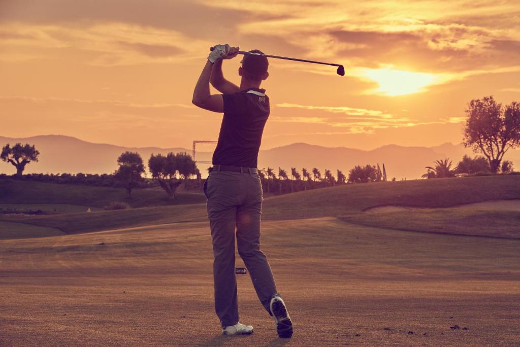 Golfa spēlētājs izsist bumbu uz fona saulrieta, kurā redzami kalni un koki. Daba un sports apvienojas, radot mierīgu atmosfēru.