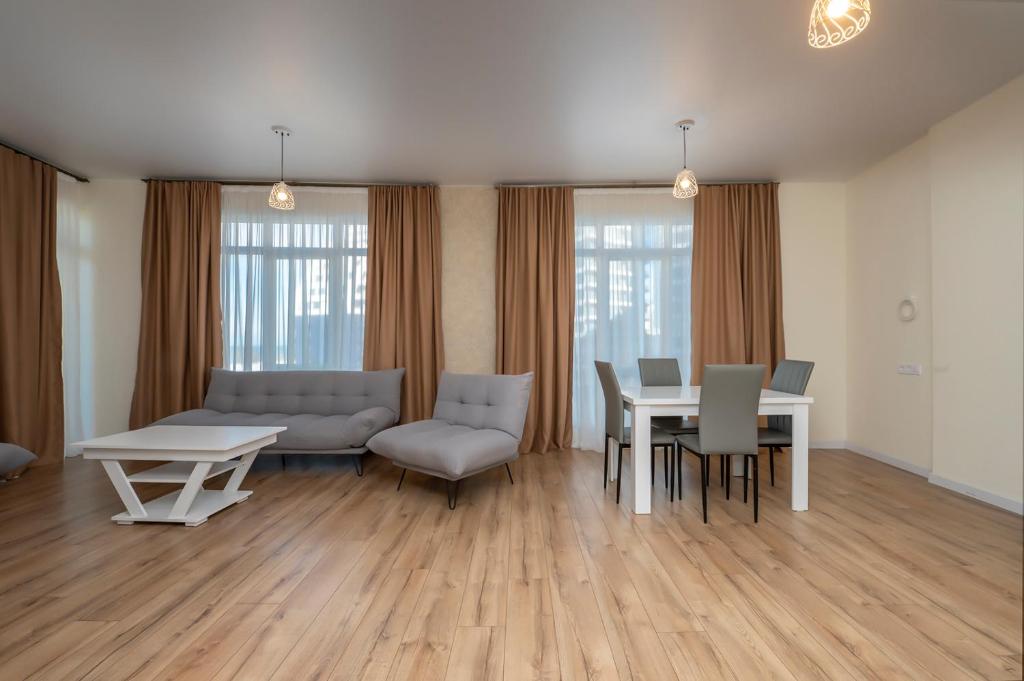Mūsdienīgs dzīvojamā istaba ar pelēkiem dīvāniem, baltu galdu un krēsliem, apgaismota ar elegantu lampu, lieli logi un siltas krāsas aizkari.