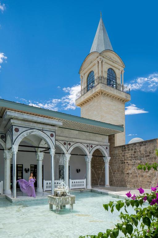 Paviljonu ar mitriem arku elementiem un minaretu, kas redzams fonā, kopā ar sievieti violetā kleitā, kas stāv pie strūklakas.