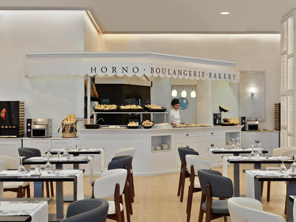 Mūsdienīga maiznīca ar elegantu dizainu, kurā redzama pavāre, kas strādā pie maizes un konditorejas izstrādājumiem. Gaisīga interjers ar baltām un koka mēbelēm, kas rada patīkamu atmosfēru.