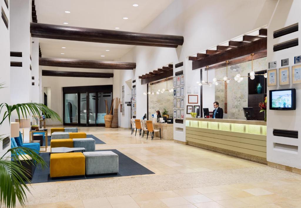 Moderns viesnīcas reģistratūras interjers ar gaišām sienām, koka griestu sijām un krāsainām mēbelēm. Viesnīcas darbinieks apkalpo viesus pie reģistratūras.