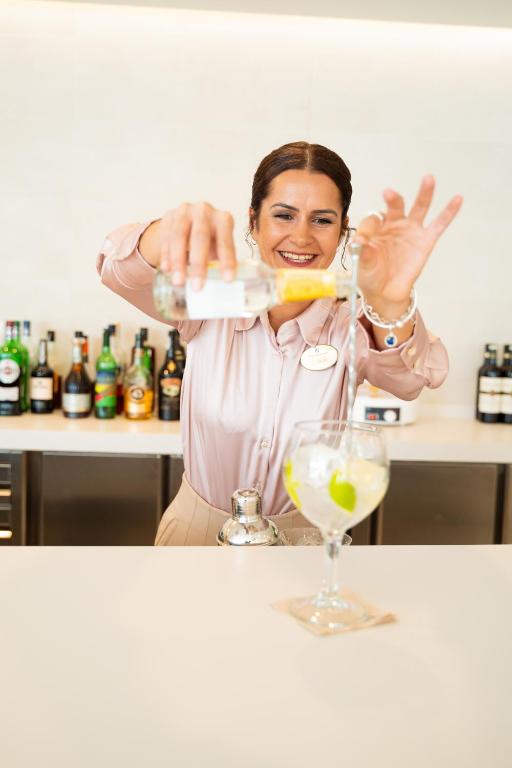 Barmene sajauc dzērienu, pievienojot citronu un laimu, ar prieku pasniedzot to klientiem. Fonā redzami dažādi alkoholiskie dzērieni uz bāra.