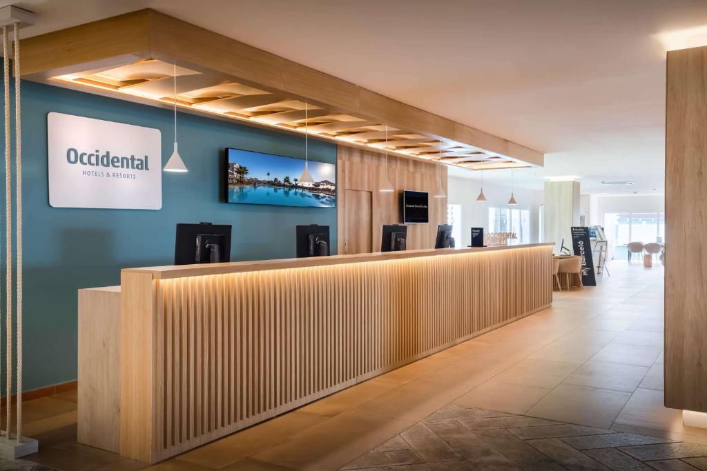 Mūsdienīga viesnīcas reģistratūra ar koka apdari un gaismas elementiem, kurā redzama "Occidental Hotels & Resorts" zīme un televizors, kas rāda viesnīcas teritoriju.