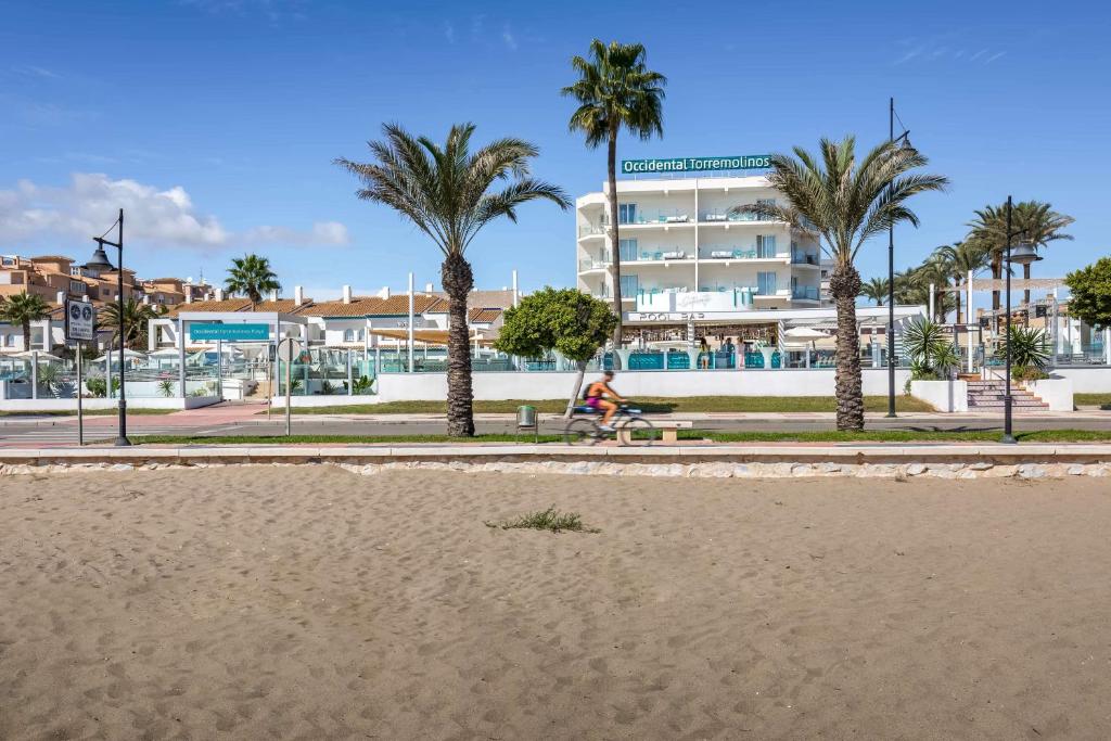 Pludmales ainava ar palmām un viesnīcu "Occidental Torremolinos", kas atrodas pie ūdens. Ceļotājs brauc ar velosipēdu gar promenādi.