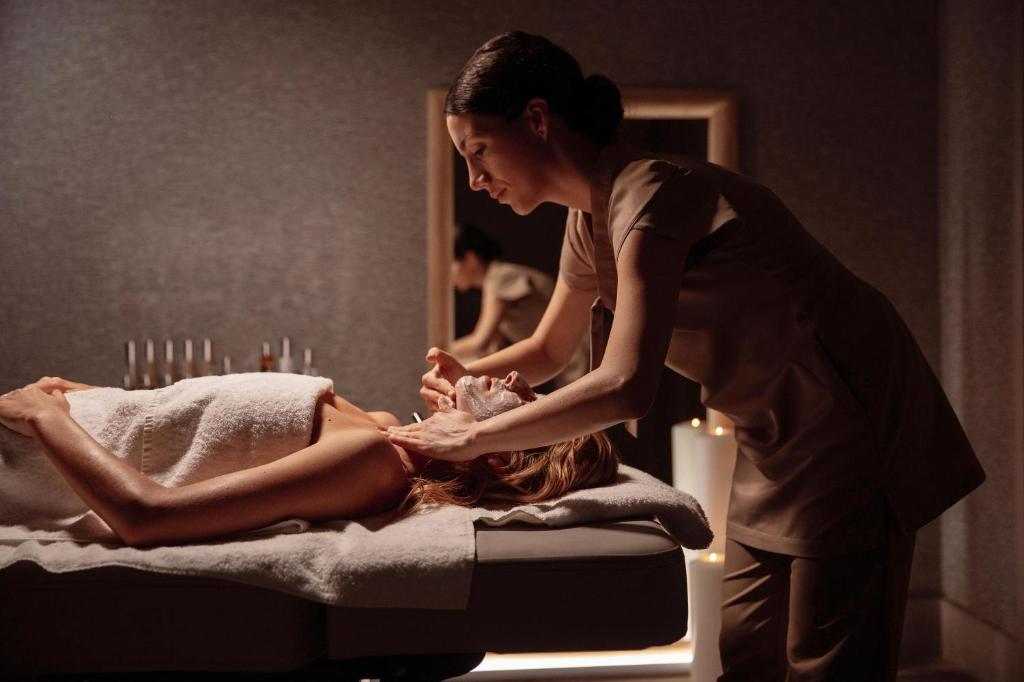Sieviete saņem relaksējošu sejas masāžu spa centrā, kamēr otra sieviete profesionāli veic procedūru. Apkārt ir mierīga atmosfēra ar svečiem un relaksējošu apgaismojumu.