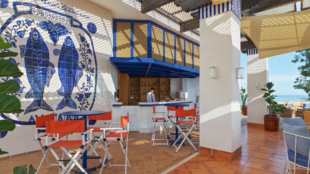 Moderns bārs ar zilām un baltām flīzēm, kas attēlo zivis, un ērtām sēdvietām. Skats uz pludmali un sauli caur atvērto telpu.