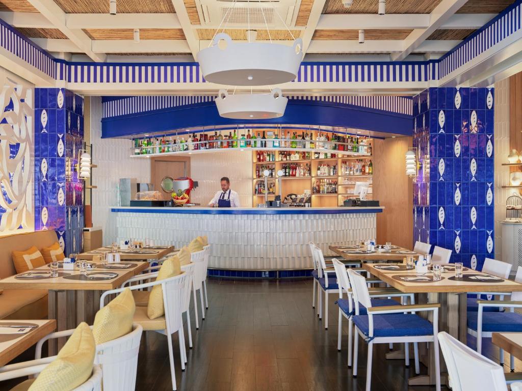 Mūsdienīgs restorāns ar zilas un baltas krāsas interjeru, kurā redzams bārmenis aiz bāra un eleganti sakārtotas galdi. Ideāla vieta relaksācijai un gastronomiskām baudām.