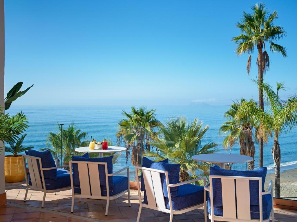 Jūras skats no terases ar ziliem krēsliem un galdiem, apkārt palmām un saulainu debesu fonu. Ideāla vieta relaksācijai pie ūdens.