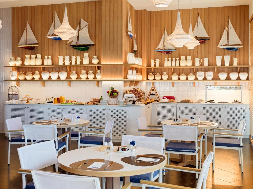 Mūsdienīgs restorāns ar baltām mēbelēm un jūras tematiku, kurā redzami burājošu laivu dekori un stilīgi trauki uz plauktiem. Gaisma ir silta un patīkama, radot viesmīlīgu atmosfēru.