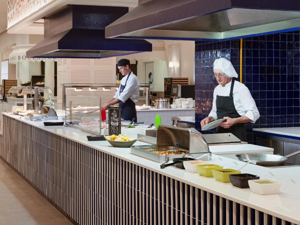 Virtuves darbinieki strādā pie modernās bufetes, gatavojot ēdienus un organizējot sastāvdaļas. Skats uz tīru un sakārtotu virtuvi, kurā tiek piedāvāta dažāda veida pārtika.