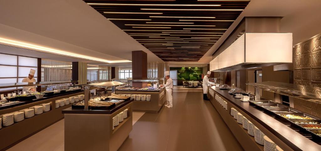 Mūsdienīgs restorāna interjers ar bufeti, kurā redzami šefpavāri, kas gatavo ēdienus. Gaismas un tekstūras rada siltu un aicinošu atmosfēru.