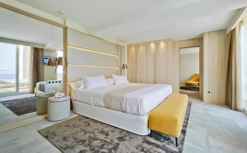 Mūsdienīgs guļamistabas interjers ar komfortablu gultu, dzeltenu banketu un plašu logu, kas piedāvā skatu uz jūru. Telpa ir gaiša un eleganta, ar moderna dizaina elementiem.