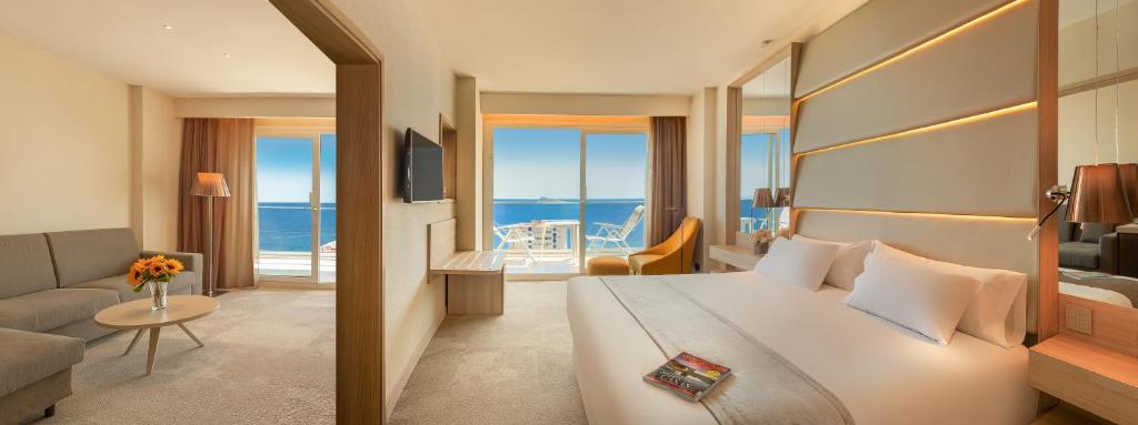 Mūsdienīgs viesnīcas numurs ar jūras skatu, kas ietver ērtu gultu, dīvānu un terasi. Interjers ir gaišs un elegants, radot relaksējošu atmosfēru.