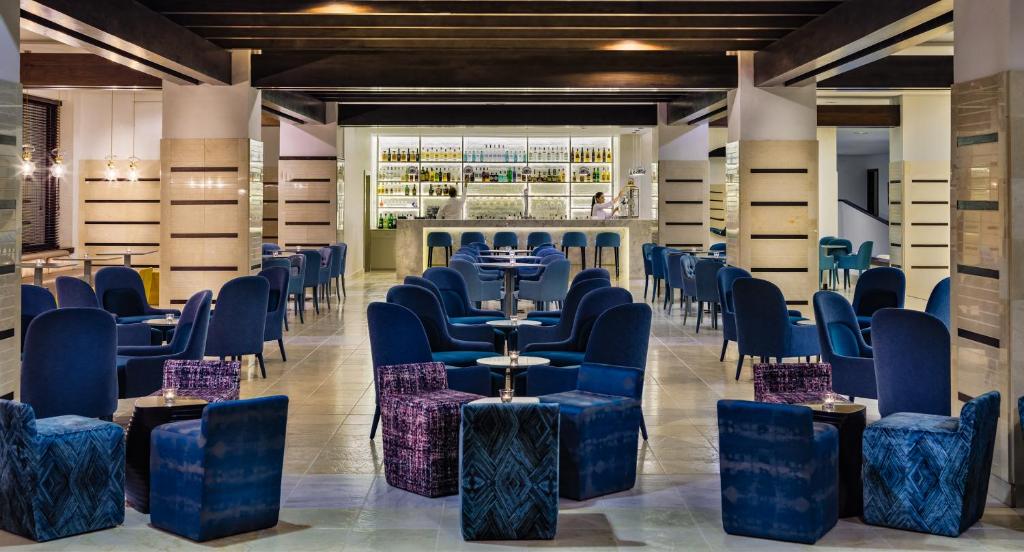 Mūsdienīgs bārs ar zilu krēslu un galdu izkārtojumu, kas atrodas gaišā telpā ar elegantu dekoru un dzērienu plauktu. Ideāla vieta relaksācijai un socializēšanai.