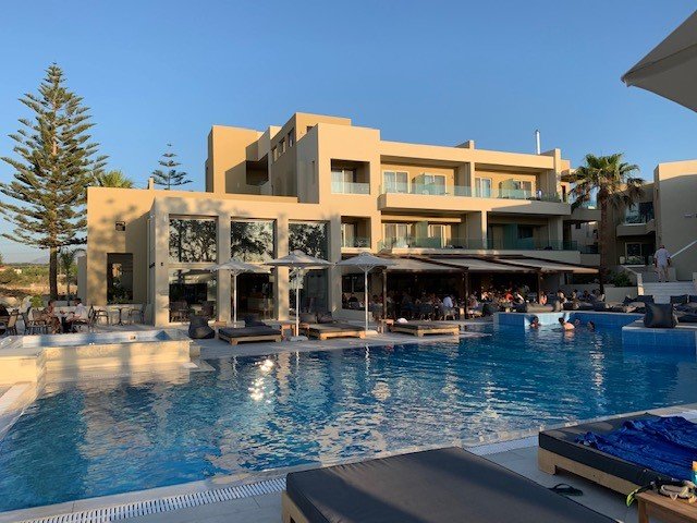 Mūsdienīga viesnīca ar plašu baseinu un terasi, kurā redzami viesi, kas atpūšas. Apkārt augstie palmas un zila debesis rada relaksējošu atmosfēru. panoramic view with beach and pools