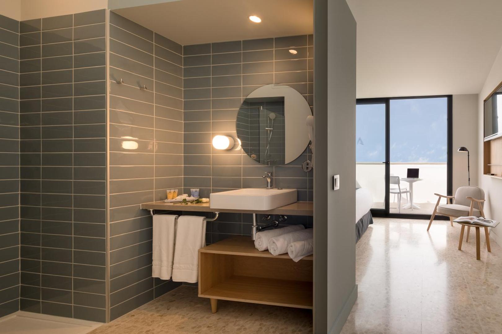 AQUA Hotel Silhouette & Spa moderns numurs ar atvērtā plānojuma vannasistabu, pelēkām flīzēm, apaļu spoguli un skatu uz balkonu Malgrat de Mar.