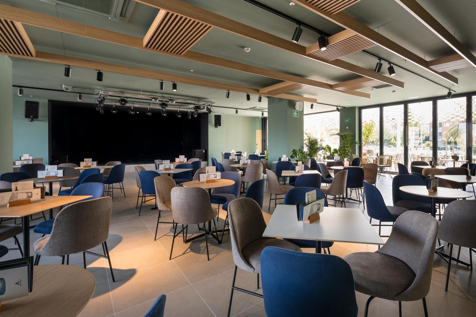 AQUA Hotel Silhouette & Spa plašā atpūtas zāle ar skatuvi pasākumiem, moderniem krēsliem un galdiņiem viesu izklaidei Malgrat de Mar kūrortā.