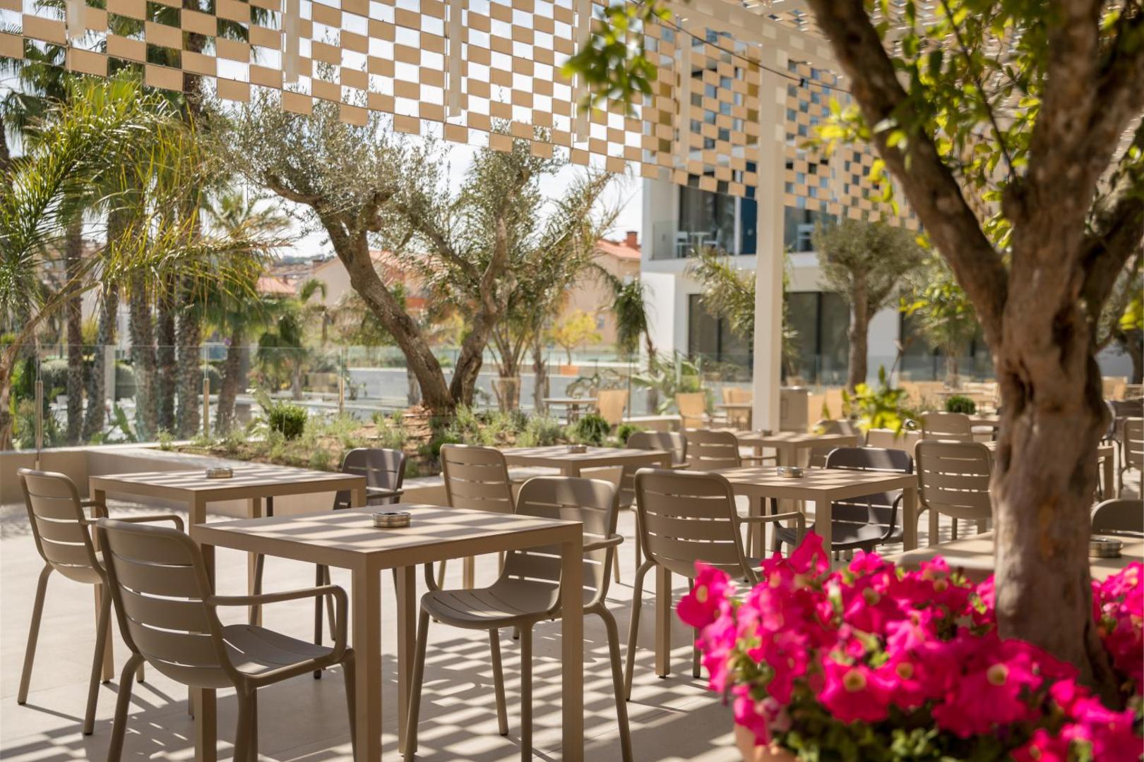 AQUA Hotel Silhouette & Spa āra terases restorāns ar galdiņiem zem dekoratīvas nojumes, ko ieskauj ziedoši augi un koki.