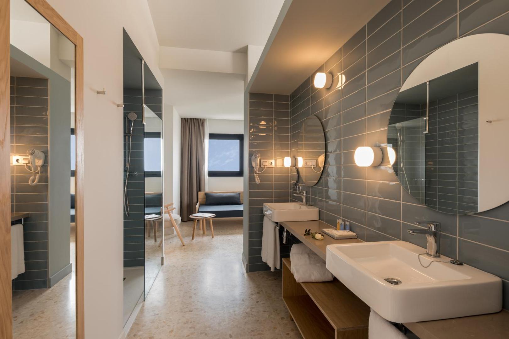 AQUA Hotel Silhouette & Spa numura vannasistaba ar modernu dizainu, pelēkām flīzēm, apaļiem spoguļiem un atvērtā plānojuma dušu.