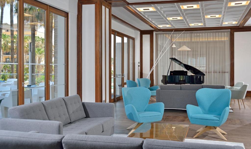 Mūsdienīgs viesnīcas interjers ar pelēku dīvānu, zilu krēslu grupu un melnu pianīnu, kas nodrošina elegantu atmosfēru. Plašas logu ailes ļauj ienākt dabiskai gaismai un sniedz skatu uz apkārtējiem palmām.