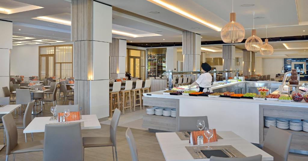 Mūsdienīgs restorāns ar gaišu interjeru, kurā redzami galds ar traukiem un bufete ar dažādiem ēdieniem. Eleganti gaismas ķermeņi un silti toņi rada patīkamu atmosfēru.