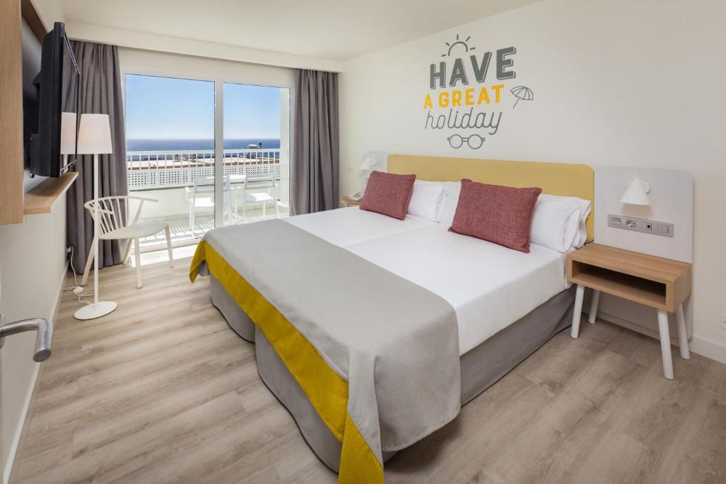 Mūsdienīgs viesnīcas numurs ar divām gultām, dekorēts ar spilgtiem akcentiem un skatu uz jūru. Sienā redzama uzraksts "Have a great holiday", kas piešķir viesnīcas atmosfērai patīkamu noskaņu.