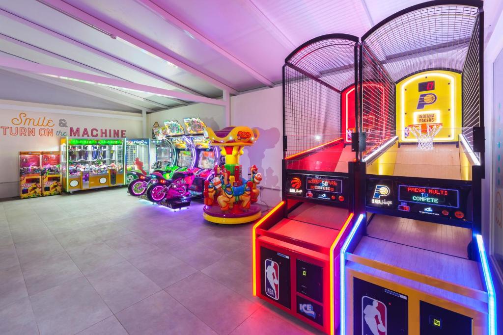 Spēļu istaba ar dažādiem arcade spēļu automātiem, tostarp basketbola spēle un izklaidējošas braucamās mašīnas. Telpa ir spilgti apgaismota un aicina apmeklētājus izbaudīt spēles.