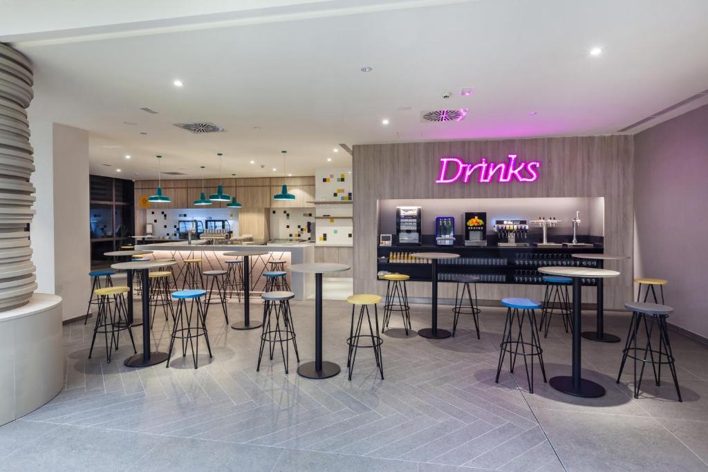 Mūsdienīgs bārs ar neonisku "Drinks" zīmi, krāsainiem krēsliem un stilīgu interjeru, ideāls vieta atpūtai un dzērienu baudīšanai.
