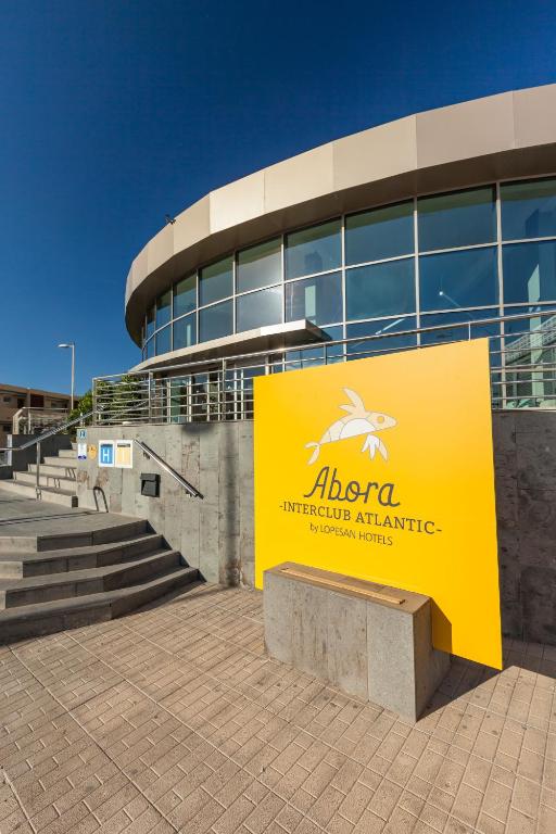 Dzeltena zīme ar nosaukumu "Abora Interclub Atlantic" pie viesnīcas ieejas, ar modernu arhitektūru un skaidru zilo debesu fonu.