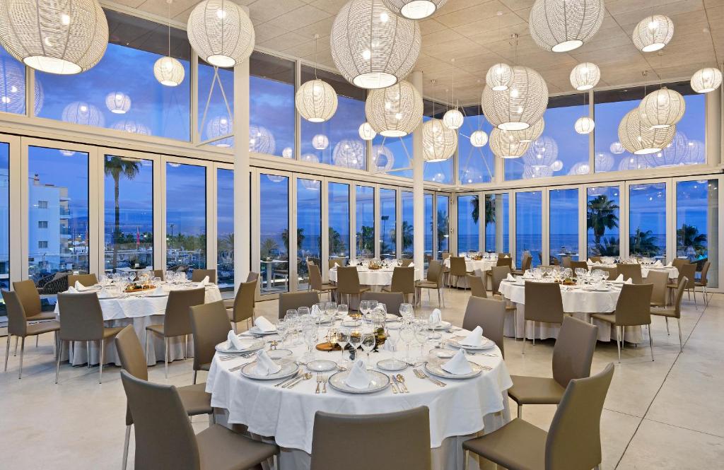 Elegantā restorāna iekšiene ar baltām galdautām un izsmalcinātiem galda piederumiem, apgaismota ar modernām lampām un ar skatu uz jūru.