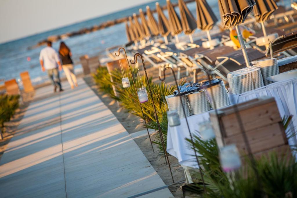 Pludmales pastaigu ceļš ar pāri, kas dodas prom, fonā ir saulriets un pludmales sauļošanās krēsli. Blakus atrodas svinību galds ar šķīvjiem un dekoratīvām lampiņām.