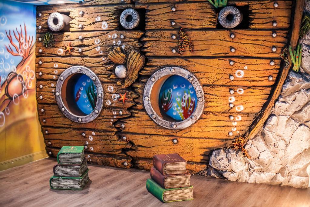 Krāsains sienas gleznojums ar divām apaļām lodziņām, kas atgādina zemūdens skatu, un kaudze ar grāmatām priekšpusē. Ideāla vide bērnu izglītības un izklaides telpām.