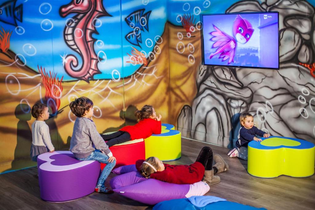 Bērni atpūšas uz krāsainām mīkstām mēbelēm, skatoties uz ekrānu, kur tiek rādīta animācija. Fons ir krāsains ar jūras dzīvniekiem un burbuļiem, radot jautru atmosfēru.