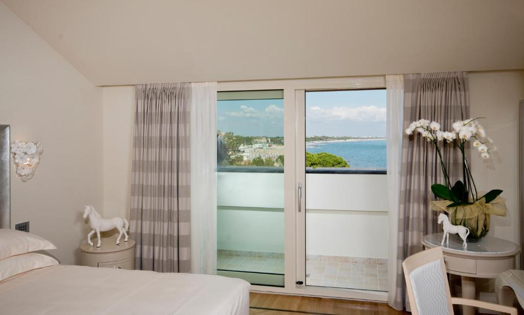Mūsdienīgs guļamistabas interjers ar skatu uz jūru, kurā redzami eleganti aksesuāri, piemēram, balta ziedu kompozīcija un dekoratīvi poniji. Gaismas tonis un logi rada patīkamu atmosfēru.