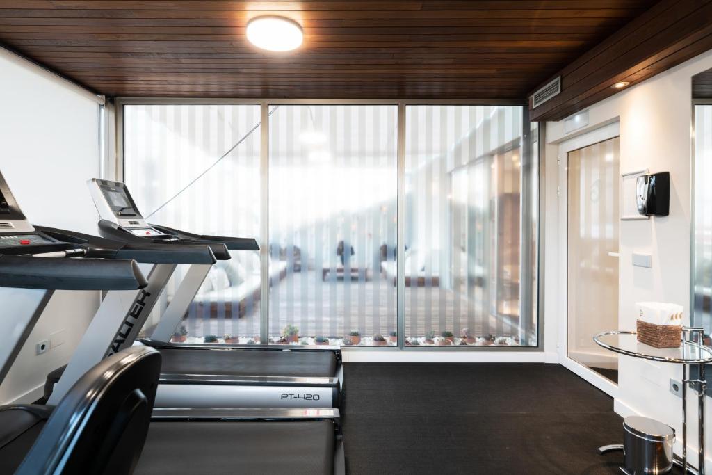 Mūsdienīga trenažieru zāle ar skatu uz ārpusi, kur redzami skrejceliņi un lieli logi ar žalūzijām. Telpa ir gaiša un eleganta ar koka griestiem.