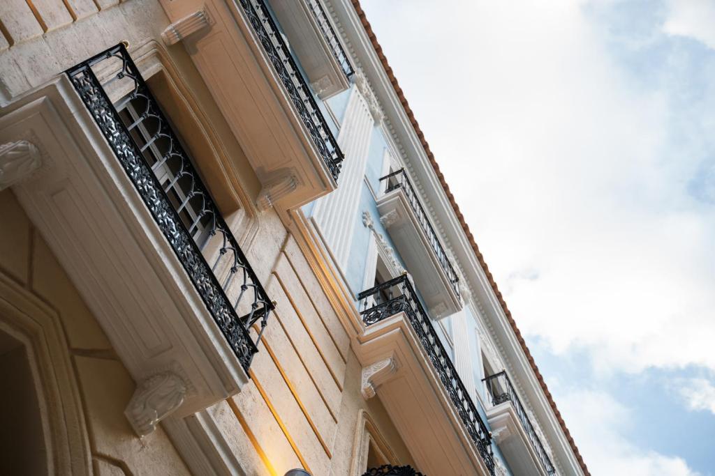 Balto ēku ar melnām balkonām skats no apakšas, redzami dekoratīvi arhitektūras elementi un debesis fonā.