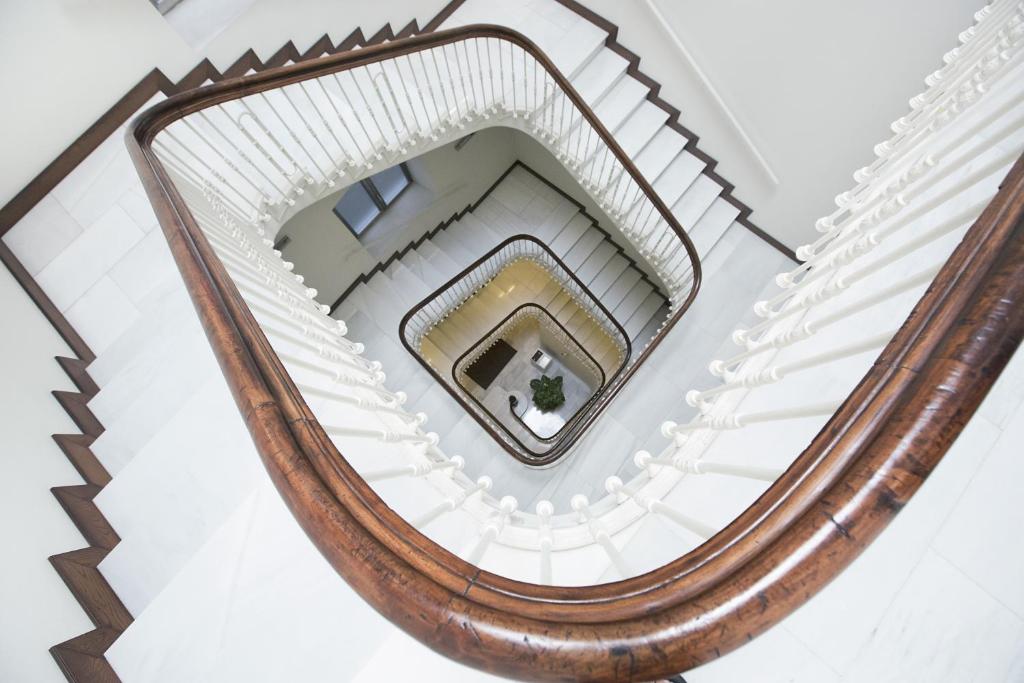 Skaista spirālveida kāpņu telpa ar koka margām un baltu flīžu grīdu, kas rada elegantu atmosfēru. Kāpnes vērstas uz leju, atklājot apakšējo stāvu ar zaļo augu.