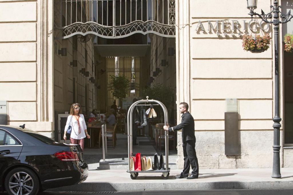 Viesnīcas portieris stāv pie iebraukšanas, stumjot bagāžas ratiņus ar somām, kamēr apmeklētāja dodas ārā. Fons iekļauj viesnīcas ēku ar nosaukumu "AMERIGO".