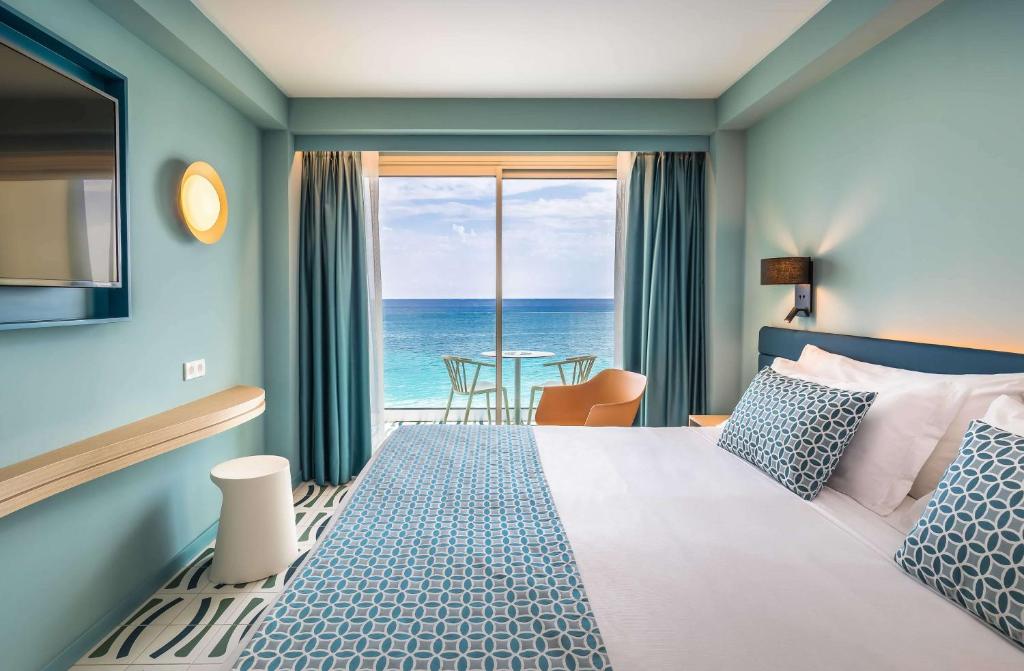 Moderns viesnīcas numurs ar skatu uz jūru, gaišām krāsām un komfortablu interjeru. Logā redzams balkons ar galdu un krēsliem, radot relaksējošu atmosfēru.