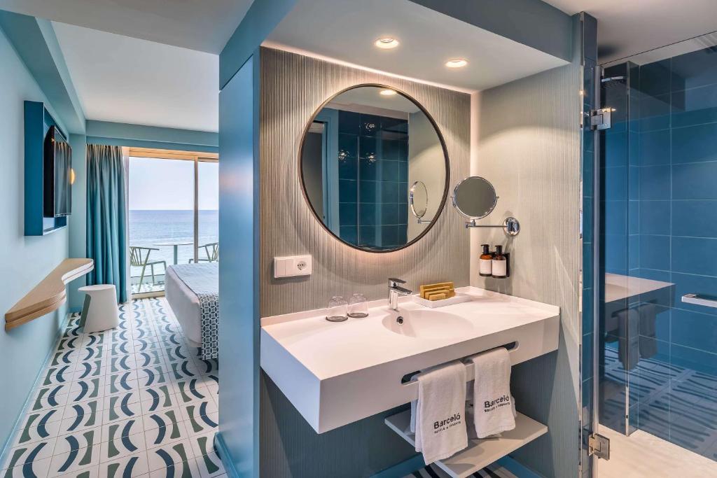 Mūsdienīga viesnīcas numura vannas istaba ar elegantu dizainu, apaļu spoguli un skatu uz jūru. Interjerā ir gaiši toņi un stilīga flīžu grīda.