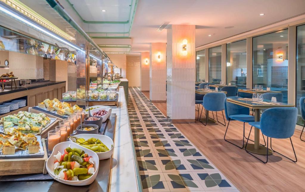 Mūsdienīga restorāna iekšiene ar bufetes stilu, kur redzami dažādi ēdieni un dzērieni, kā arī stilīgas zilas krēsli un galds. Gaismas un interjers rada patīkamu atmosfēru.