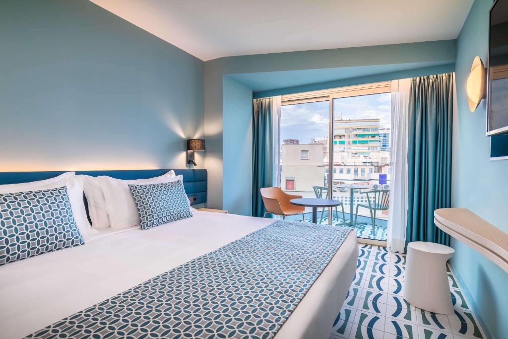 Moderns viesnīcas numurs ar gaišām krāsām, lielu logu un balkonu. Gultā ir dekoratīvas spilvendrānas, un telpā ir stilīgas mēbeles.