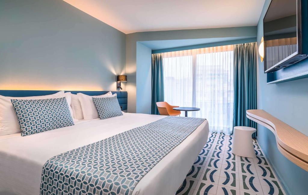 Mūsdienīgs viesnīcas numurs ar divguļamo gultu, dekorēts gaišās krāsās un ar modernu interjeru, ietverot mazu galdiņu un krēslu. Lielie logi nodrošina dabisko apgaismojumu.