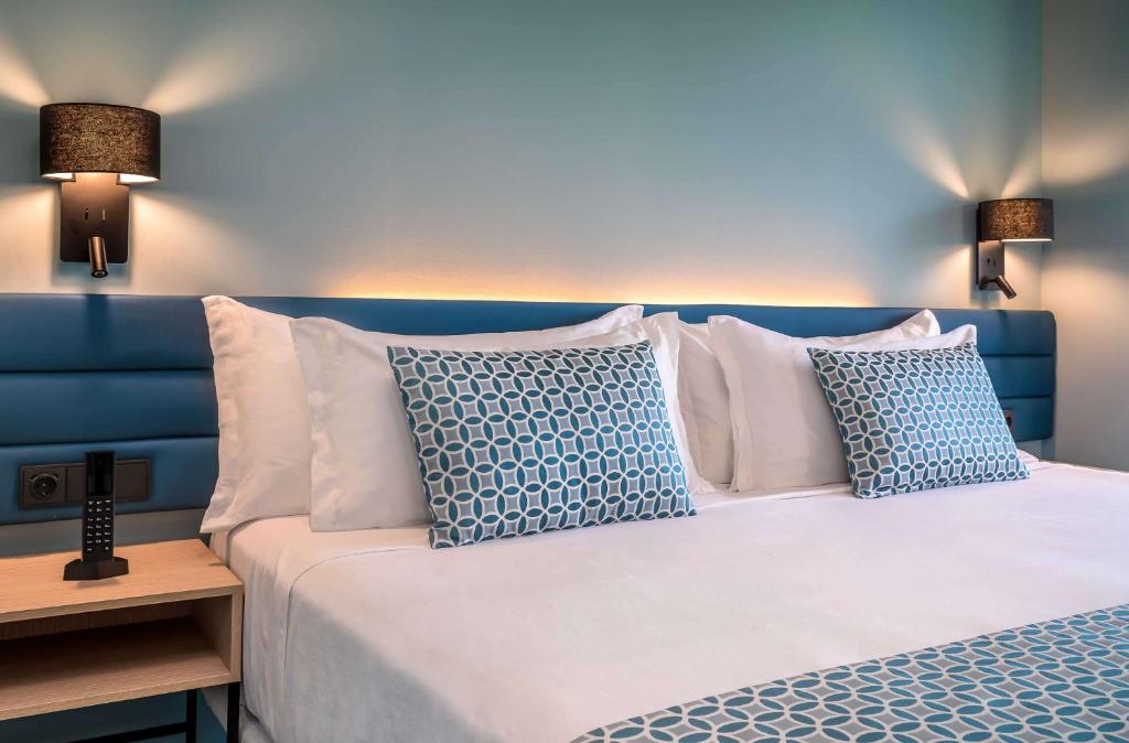 Mūsdienīga guļamistaba ar zilu sienu un gultu, kas dekorēta ar rakstainām spilveniem, apgaismota ar siltu gaismu un papildināta ar koka naktsgaldiņu un tālruņa ierīci.